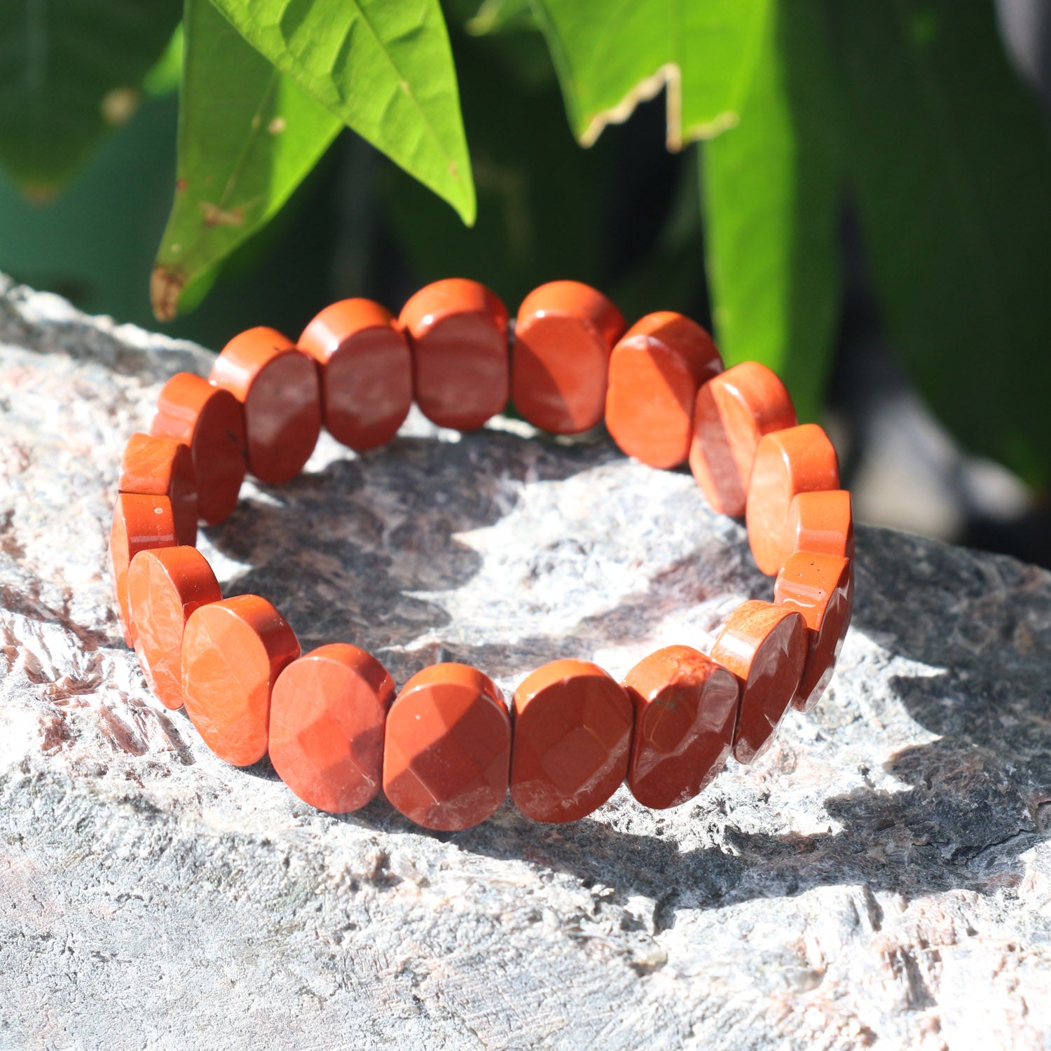 A red jasper bracelet made of polished, round, orange-red stones is displayed on a rough stone surface with green leaves in the background, brightly lit by sunlight.