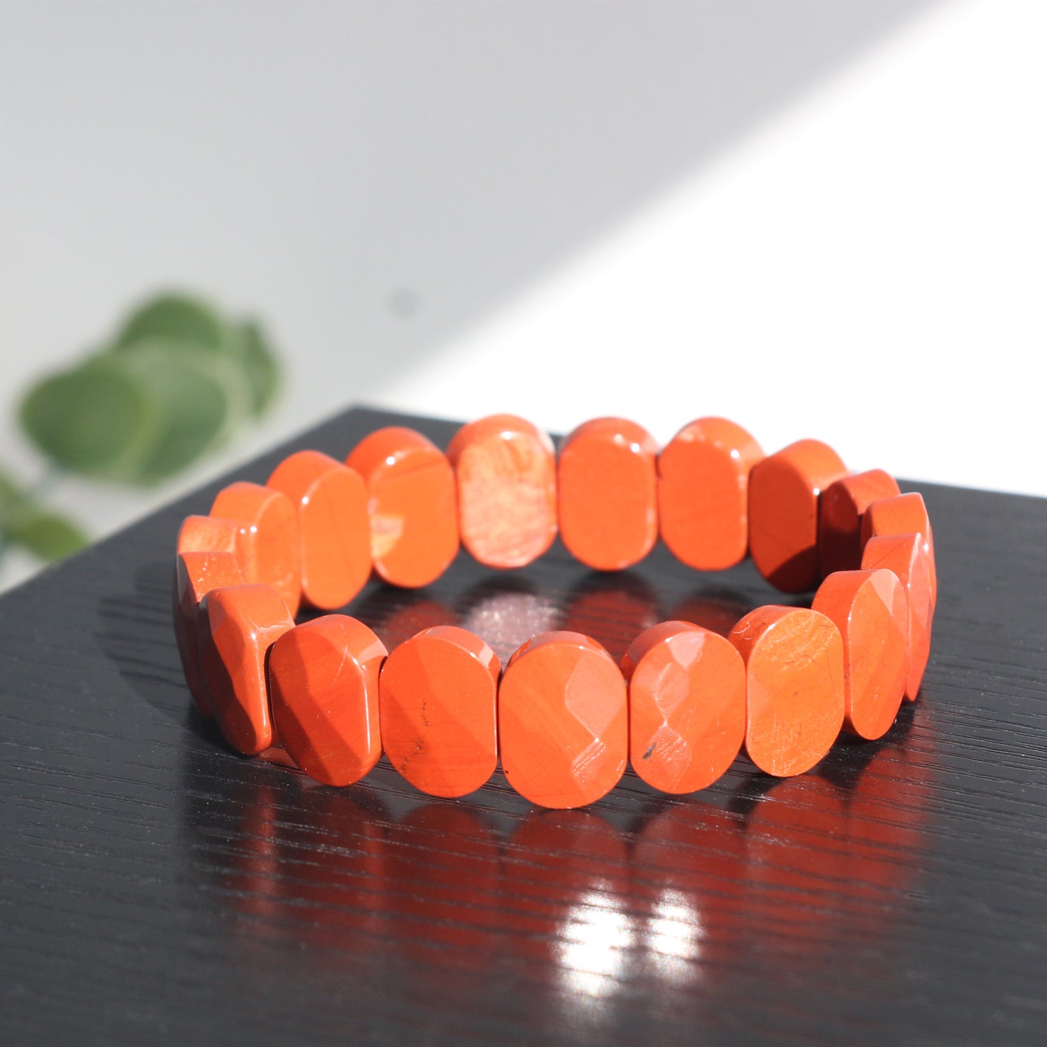 A chunky orange bracelet made of oval-shaped beads is displayed on a glossy black surface, with sunlight casting reflections and a blurred green plant in the background.